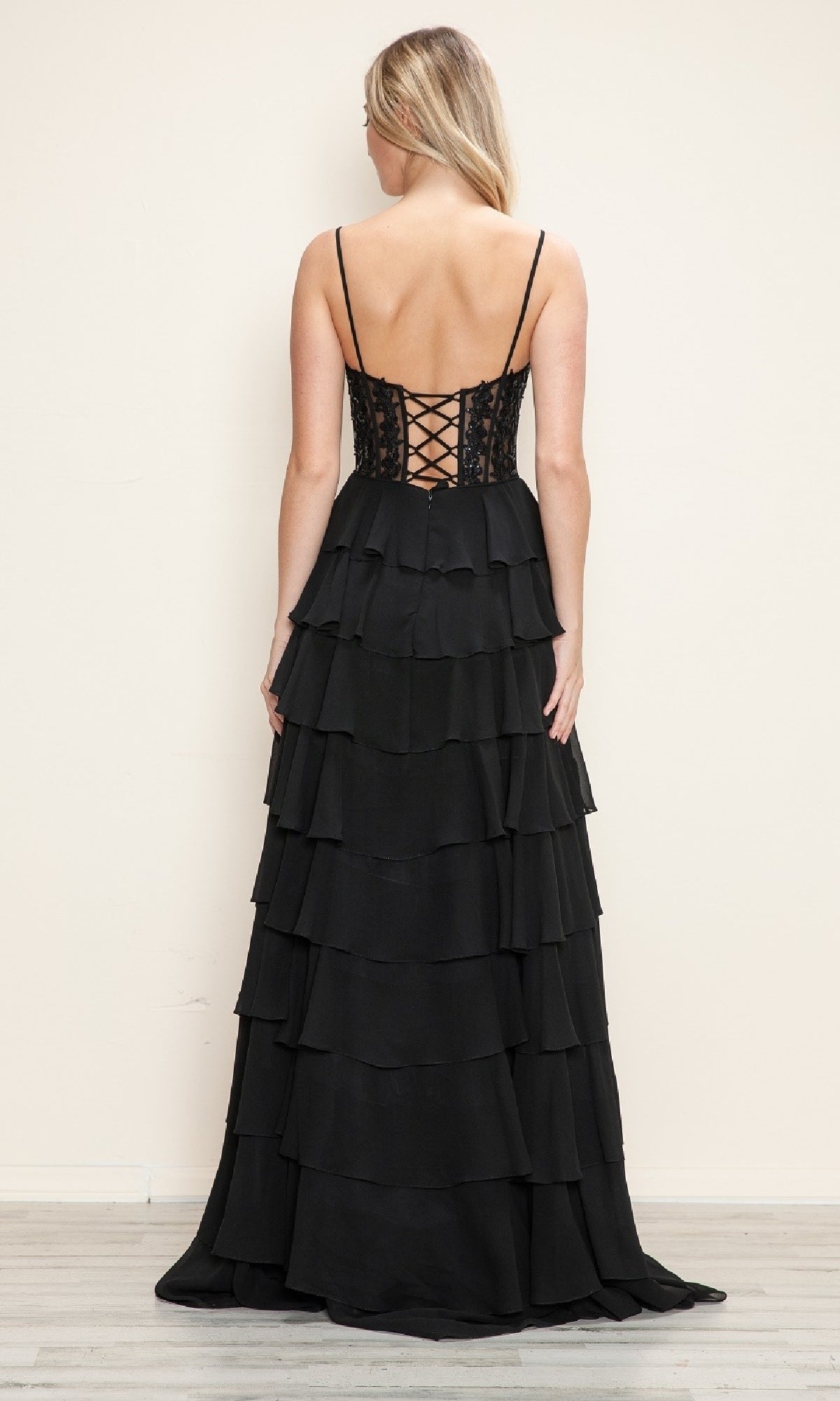A woman stands with her back to the camera, wearing the Poly USA 9698 Long Tiered Corset Prom Dress—floor-length, black, a-line with a corset-style bodice—against a plain light-colored wall.